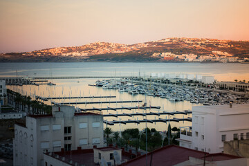 Sunset view of Tangier marina with yachts and piers, calm harbor waters, and hillside cityscape across the bay under warm evening light.