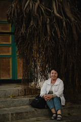 Elderly woman sitting on stone steps beneath massive hanging tree roots, calm and thoughtful portrait blending human presence with powerful natural forms.
