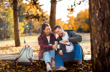 Romantic young multiracial couple enjoying a date in an autumn park, sitting on grass drinking tea.