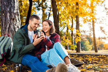 Romantic young multiracial couple enjoying a date in an autumn park, sitting on grass drinking tea.