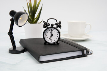 Modern business workspace with alarm clock on notebook, desk lamp, pen, coffee cup, and plant....