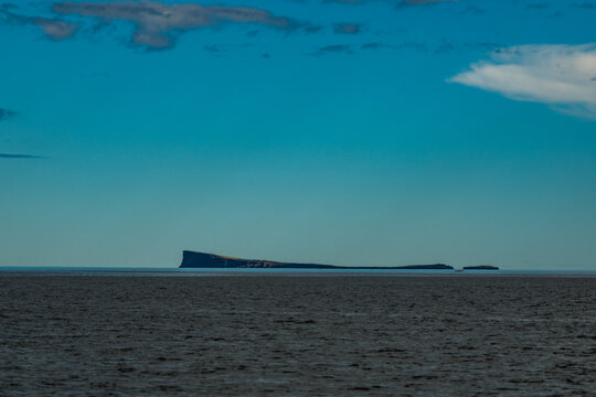 View of a distant, flat island floating on the ocean's horizon under a turquoise sky with scattered clouds, Drangey Island, Drangey Island, Iceland.