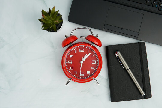 Top view office workspace with red alarm clock, notebook, pen, laptop, and plant. Concept of time management, productivity, efficiency, urgency, deadline control, and professional planning.