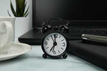 Office workspace with black alarm clock beside laptop, notebook, pen, and coffee cup. Concept of...