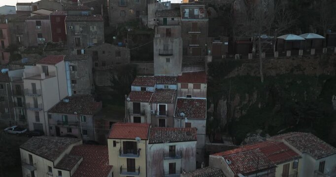 Aerial view of buildings with red and brown roofs, and a stone tower nestled into green foliage on rocky terrain, Pollina, Sicilia, Italy.