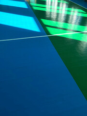 Colorful sports court floor featuring blue and green sections with bright sunlight casting geometric shadows, creating an energetic and dynamic atmosphere for athletic activities