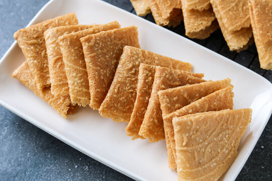 Closeup of crispy, golden-brown Kue Semprong or Love Letter cookies, a thin, folded Southeast Asian snack. Arranged on a white plate and a cooling rack, highlighting their delicate texture.