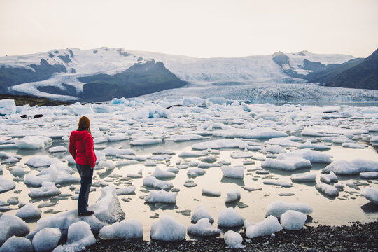 View of a person in a red jacket standing amidst the icy expanse of Fjallsarlon glacial lagoon, as icebergs drift towards the distant glacier, Vatnajokull, Iceland.