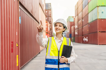 A professional female logistics manager in a hard hat and safety vest using a digital tablet and pointing while working at a busy container terminal port, overseeing international shipping trade.