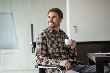 Positively smiling man in plaid shirt having coffee while sitting in the office kitchen
