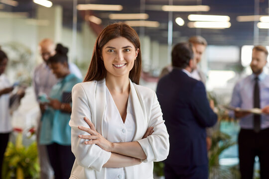 Portrait of young female company employee in conference room with arms crossed. Cheerful business woman in jacket standing after corporate training or business meeting, looking at camera and smiling.