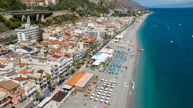 Aerial panorama of a coastal city. It is Letojanni beach, in Sicily, Italy. Colorful buildings and organized beach clubs line the shore. At the horizon, in background, it's the blue Mediterranean sea.