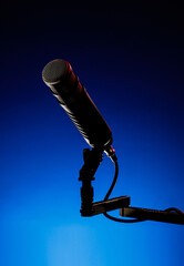 A microphone is positioned on a stand against a blue background. The setting suggests it is in a recording studio during evening hours. The scene focuses on the microphone ready for use.