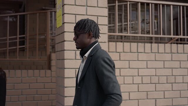 Woman intercepts man near brick building, shows phone while both hurry toward entrance, He walks with sunglasses and coat, she signals urgency over schedule, sunlight framing railing and lattice