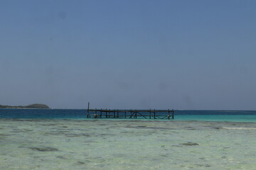 Fototapeta premium A tropical beach features a wooden pier stretching over turquoise ocean waves toward a blue horizon under a summer sky in this paradise island vacation landscape