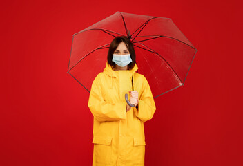 Protection concept. Portrait of young lady wearing raincoat and face surgical mask standing under umbrella, isolated over red studio background
