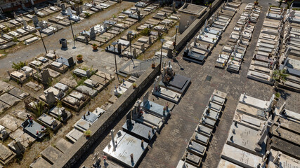 Aerial top-down view of a christian catholic cemetery. Rows of marble and granite tombs are neatly organized in blocks, separated by narrow stone paths under bright daylight.