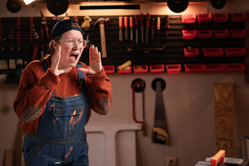 Woman joiner wears workwear in workshop scene with tools hanging on wall screaming announcement. female carpenter late evening in authentic garage.