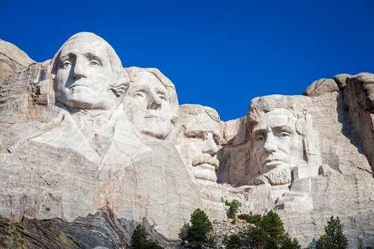 Granite carvings of Washington, Jefferson, Roosevelt, and Lincoln at Mount Rushmore National Memorial rise above pines under a clear blue sky in the Black Hills of South Dakota.