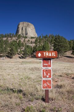 A wooden trail sign with hiking regulations stands in front of Devil's Tower National Monument in Wyoming, surrounded by grassland and pine trees beneath a clear blue sky.