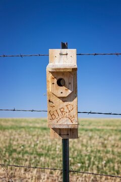 Close-up of a handmade wooden birdhouse mounted on a rural fence post with barbed wire, set against a wide open field and blue sky in Wyoming.