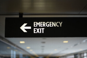 Emergency exit sign with white arrow pointing left on black background in a blurred indoor setting with ceiling lights
