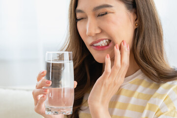 young Asian woman showing a painful facial expression and touching her cheek due to sensitive teeth or a toothache while holding a glass of water at home.