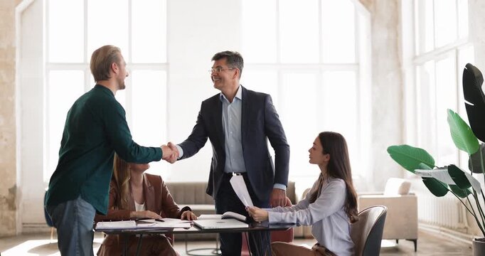 Businessman shaking hands with male client after reviewing documents, team members sit at table with reports, representing successful negotiation, partnership agreement or contract approval in office
