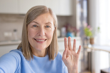 Senior woman looking at camera and waving hand, making a video call or taking a selfie in her home kitchen, connecting with family and friends through modern technology