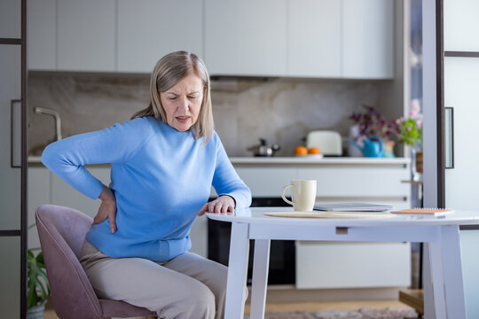 Senior woman sitting at her kitchen table, wincing and holding her lower back in pain, illustrating chronic back or kidney discomfort, aging, fatigue and home care struggles