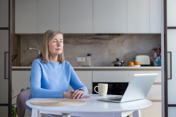 Senior woman relaxing with closed eyes at a table in a modern kitchen, practicing mindfulness and...