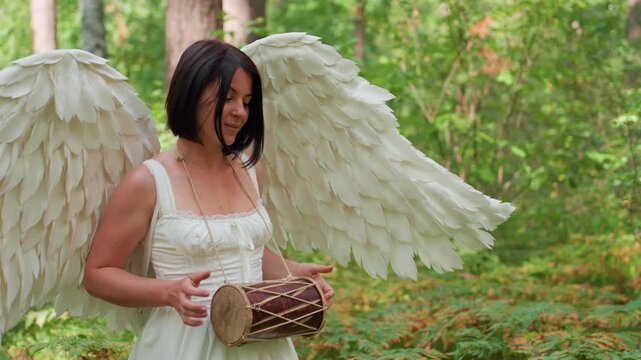 Close up of girl with white angel wings walking through green forest while gently beating small drum, sunlight filtering through leaves, peaceful rhythm echoing nature