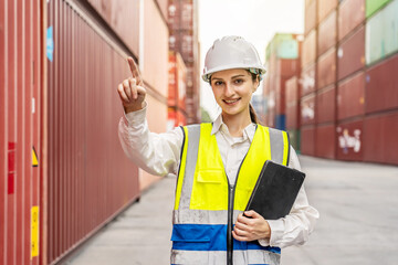 A professional female logistics manager in a hard hat and safety vest using a digital tablet and pointing while working at a busy container terminal port, overseeing international shipping trade.