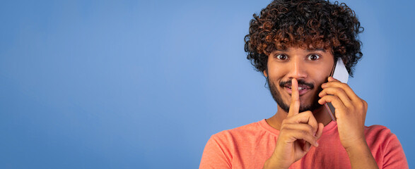 A man is holding a phone to his ear and signaling with his finger in front of his mouth. He is smiling and looking directly at the viewer. The background is blue.
