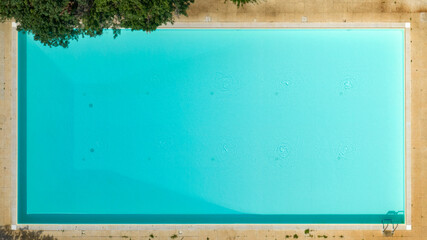 Top-down aerial view of a pristine rectangular swimming pool. The turquoise water is perfectly still, framed by a light border and a touch of green foliage in the corner. Perpendicular shot. 