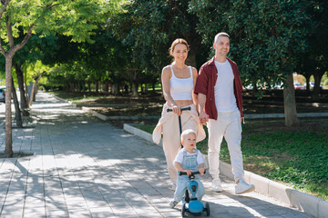 Family enjoying summer walk in park with toddler riding scooter
