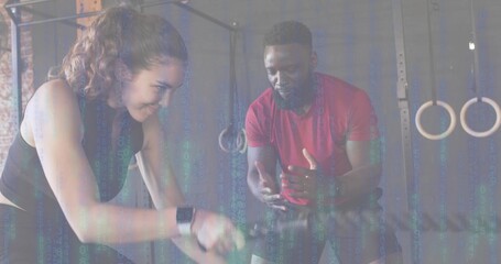 Pedaling woman in black tank gripping air bike handles at gym, coach gesturing in red shirt