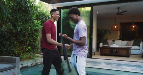 Smiling two adult men wearing maroon and purple tees exchanging green duffel bag at backyard pool