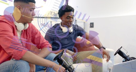 Playing two men holding controllers on sofa at home, one coral, other navy polo, with laptop