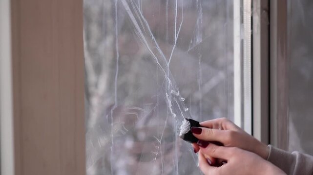 woman cleans dirty window from old scotch tape with a special tool with a blade. cleaning, tidying up, washing windows. protecting window against shock wave