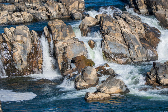 View of cascading water flowing with force and energy over jagged rocks, creating a dynamic interplay of light and shadow, Great Falls, Virginia, United States.