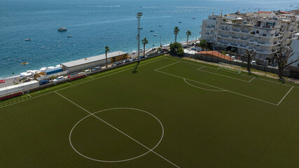 A striking aerial view of a deep green soccer field positioned just meters away from the turquoise and cobalt blue sea, separated only by a narrow pebble beach. It is a sunny summer morning. © Stefano Tammaro