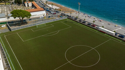 A striking aerial view of a deep green soccer field positioned just meters away from the turquoise and cobalt blue sea, separated only by a narrow pebble beach. It is a sunny summer morning.