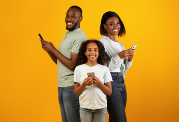 Modern family. Excited african american father, mother and daughter using smartphones while standing over yellow studio background and smiling to camera