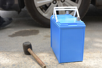 New blue car battery and rubber mallet on the ground prepared for vehicle maintenance or replacement near a car wheel.