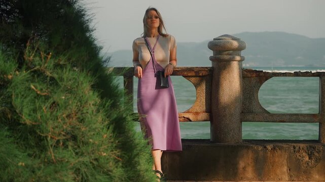 young woman, stands on a parapet in a purple dress and looks into the distance. A blonde girl looks into the distance at an island.