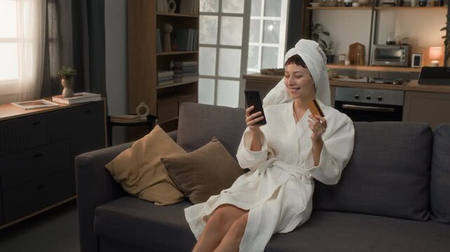 Zoom in shot of young biracial girl in bathrobe and eye patches sitting on couch at home holding smartphone and credit card and expressing joy after managing to catch great offer