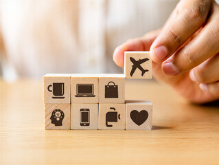 Hand placing wooden block with airplane icon on top of stack of blocks with various travel and technology symbols on light brown wood surface