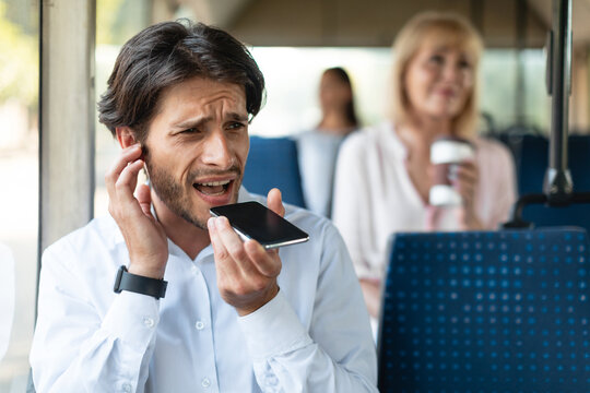 Weak Cellular Signal Concept. Upset business man talking on cellphone and plugging ear with finger sitting in public transport, angry young guy can't hear because of noise and loud passengers
