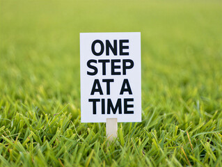 White sign on green grass with one step at a time text for motivation and inspiration in a natural outdoor setting